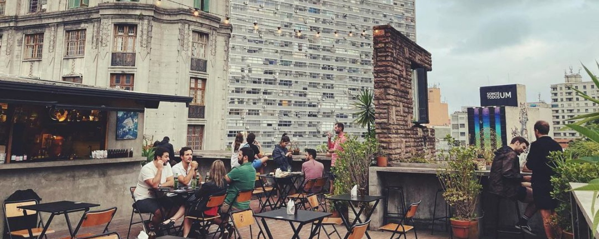 Foto de um dos rooftops em São Paulo mais conhecidos, A Balsa, com pessoas sentadas na mesa e outras conversando entre si. 