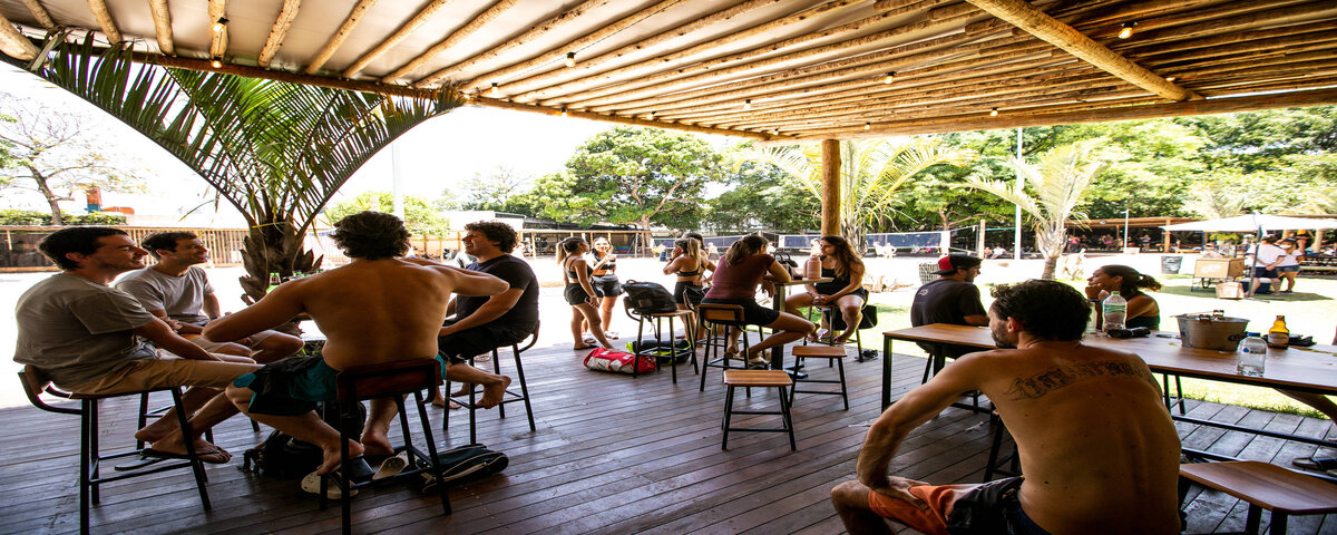 Espaço interno do Praia Brava Sand Club, com pessoas sentadas em diversas cadeiras e com bebidas nas mesas. 