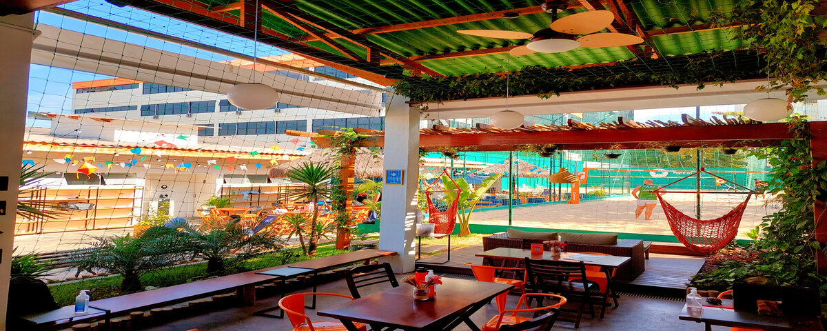 Espaço interno do Let’s Poke Praia Bar, com redes para descanso e diversas mesas e cadeiras para drinks e bebidas. 