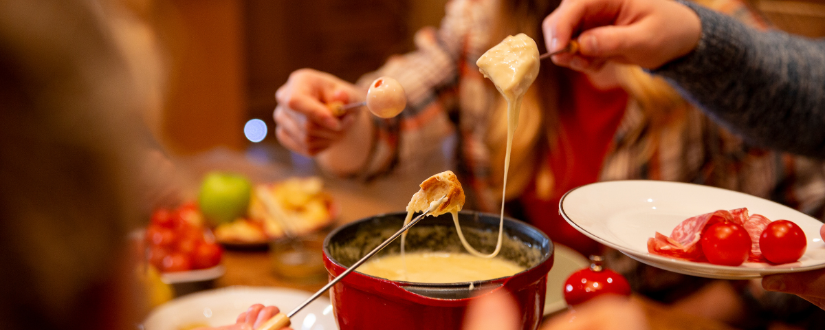 Mãos segurando um garfo espetando alimentos para misturar no fondue de queijo em cima da mesa.