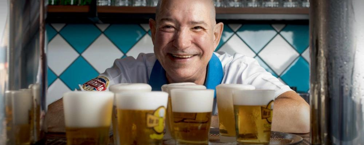Foto do dono do bar Seu Juarez, que sorri, com copos de chope em sua frente.