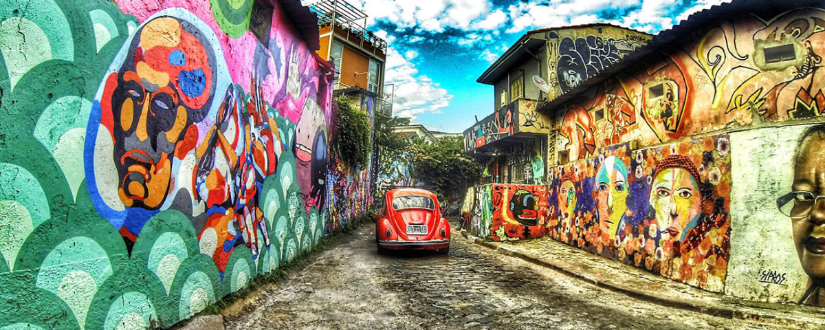 Foto do Beco do Batman, localizado na Vila Madalena, sendo um dos lugares para fotografar no Dia Mundial da Fotografia, com grafites coloridos nas paredes e um fusca vermelho no centro da imagem.