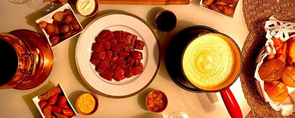 Foto de cima de uma mesa, com uma panela de queijo, cesta de pães e pratos em volta com alimentos para misturar.