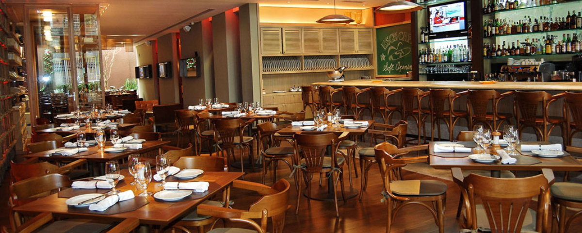 Interior do Esch Café, com cadeiras, mesas, copos, talheres e guardanapos em cima dela, tendo um bar no canto direito da imagem.