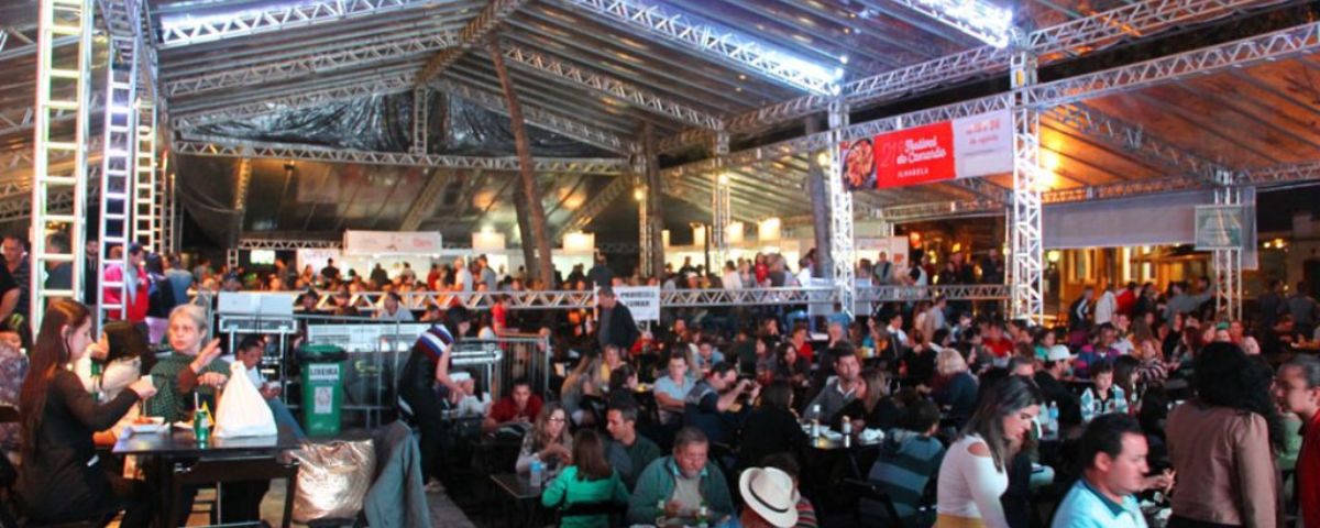 Foto de uma das edições do Festival do Camarão de Ilhabela, com muitas pessoas sentadas em mesas, outras em pé, com bebidas e comidas.