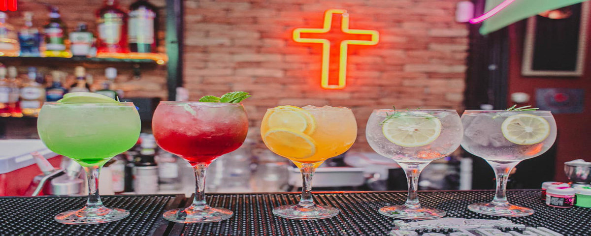 Copos com drinks um do lado do outro em cima de um balcão com um fundo de tijolinhos e uma cruz em neon vermelho.