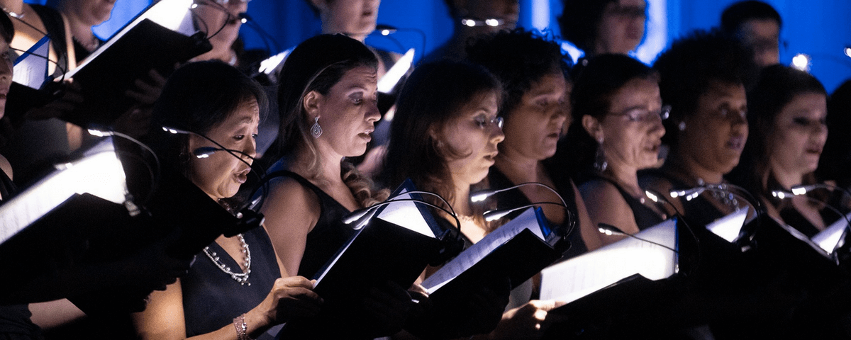 Mulheres cantando uma ao lado da outra, em uma apresentação em coro. 