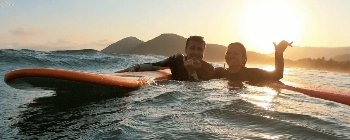 Homem e mulher no mar, com pequenas ondas, apoiados em pranchas de stand up paddle, com sol ao fundo fazendo reflexo na água.