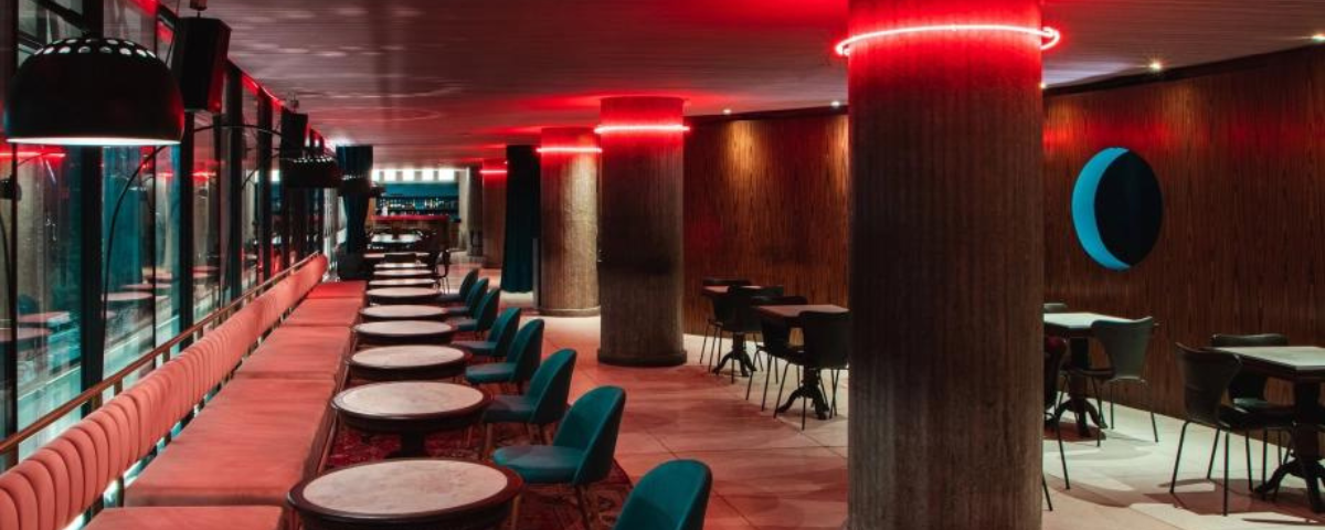 Foto do interior do Riviera bar, um dos bares abertos na segunda-feira, com cadeiras, poltronas e mesas, num ambiente com luzes vermelhas.