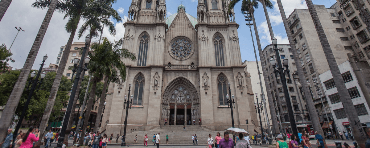 Foto de frente da Catedral da Sé, localizada no Marco Zero de São Paulo, com pessoas na rua, edifícios ao fundo e árvores pelas laterais da foto.