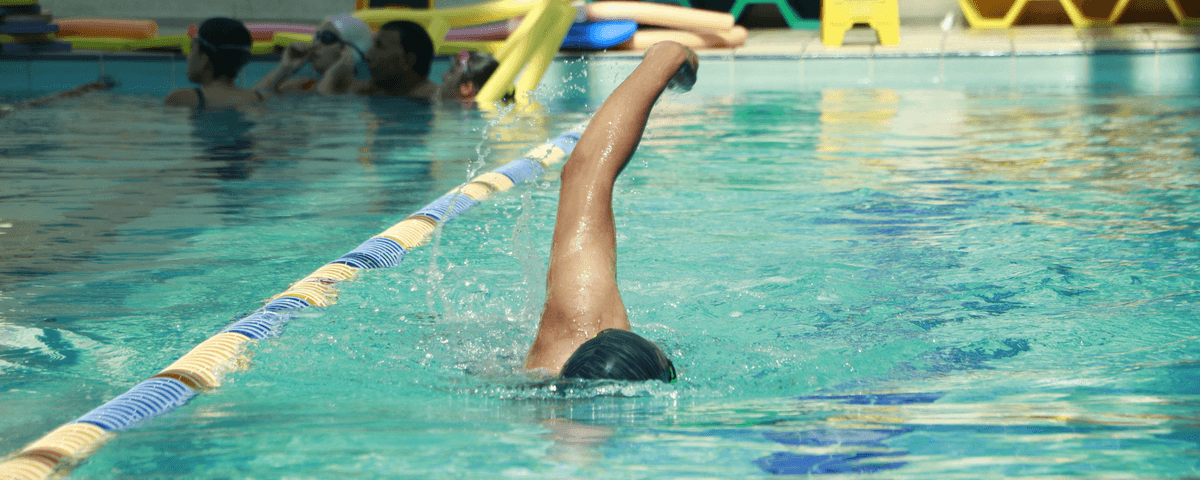 Pessoa dentro de uma piscina azul, praticando natação com o nado estilo crawl, com pessoas ao fundo. 