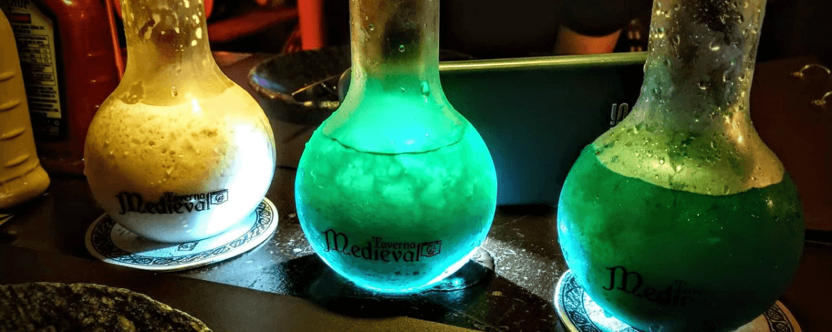 Três garrafas de bebidas no formato de um frasco erlenmeyer com líquidos nas cores verde e branco. As bebidas são da Taverna Medieval, um dos lugares geek para se conhecer em São Paulo.