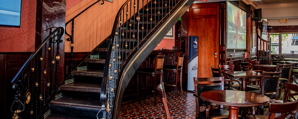 Interior do Blue Pub, um dos lugares para se tomar whisky em São Paulo. Na foto há uma escada na lateral esquerda e mesas na direita.