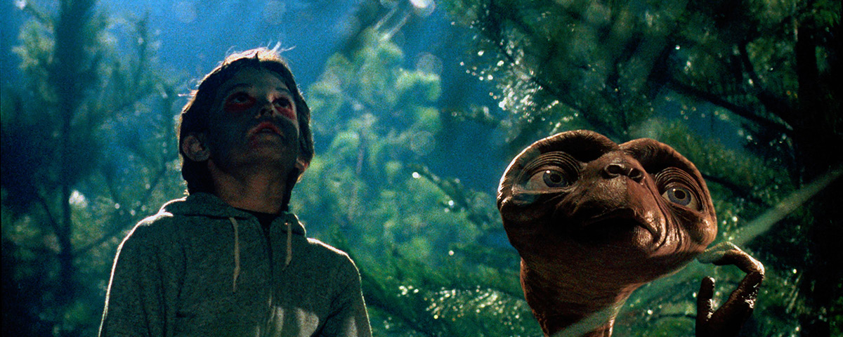 Menino e personagem ET em meio a uma floresta, com sombra reproduzida em seus rostos, olhando para cima.