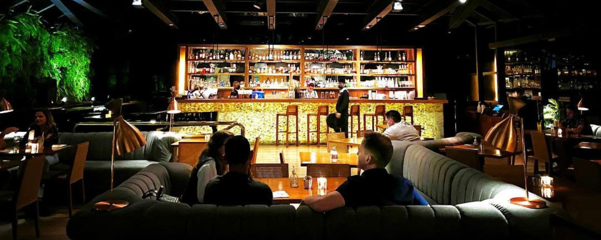 Interior do Must Bar, com pessoas sentadas ao sofá e um bar ao fundo da imagem, com barmans fazendo bebidas. 