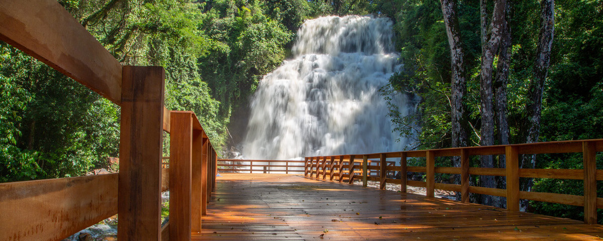 Ponte de madeira da cachoeira Cascata, com ela ao fundo, além de árvores ao redor.