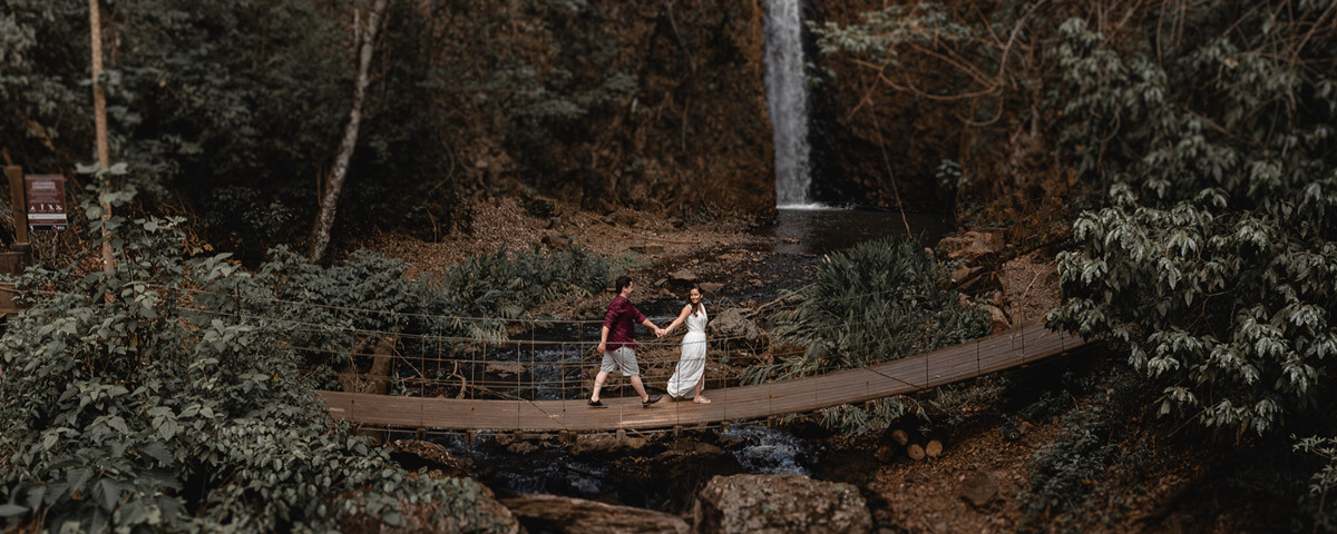 Homem e mulher de mãos dadas, andando pela ponte da cachoeira de Cassorova, cercada por árvores e vegetação.