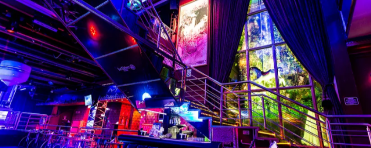 Interior da balada Le Rêve Club, com luzes em neon azul e vermelho, escada, balcão e bebidas sobre ele. 