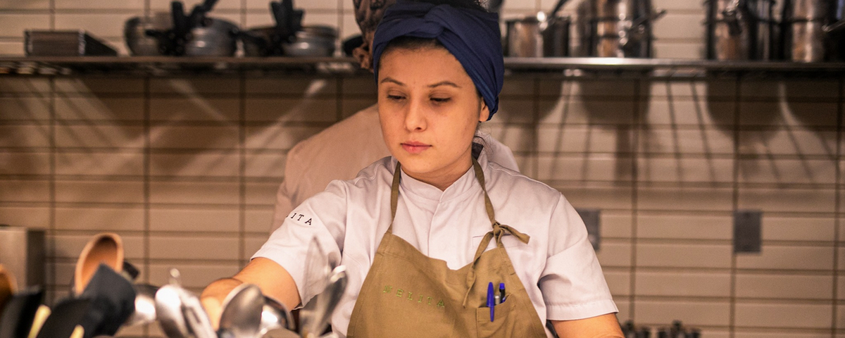 Cozinheira do Nelita Restaurant, usando um avental bege, com dolmã branco. 