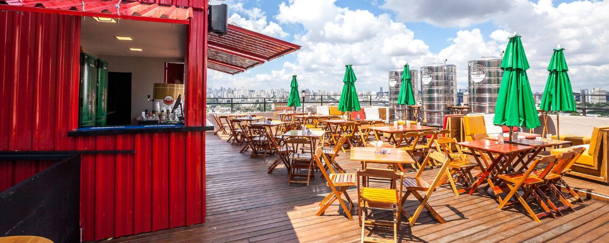 Área externa do Oh Freguês, um dos bares para ir com os amigos em São Paulo, com mesas e cadeiras de madeira, guarda-sol verde sobre as mesas e um bar vermelho na lateral esquerda da imagem.
