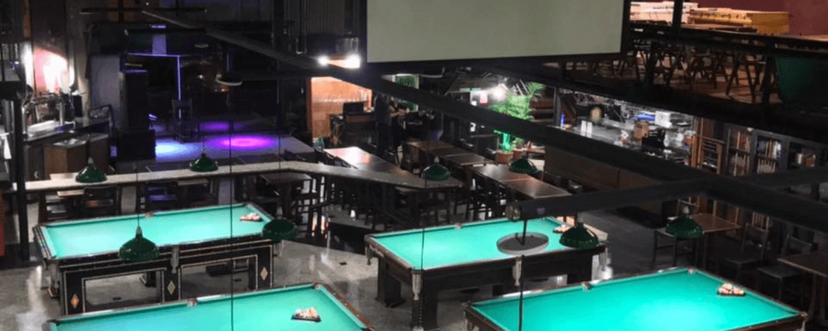 Interior do Dona Mathilde Snooker Bar, com mesas de sinuca, um telão e um bar à direita da imagem.