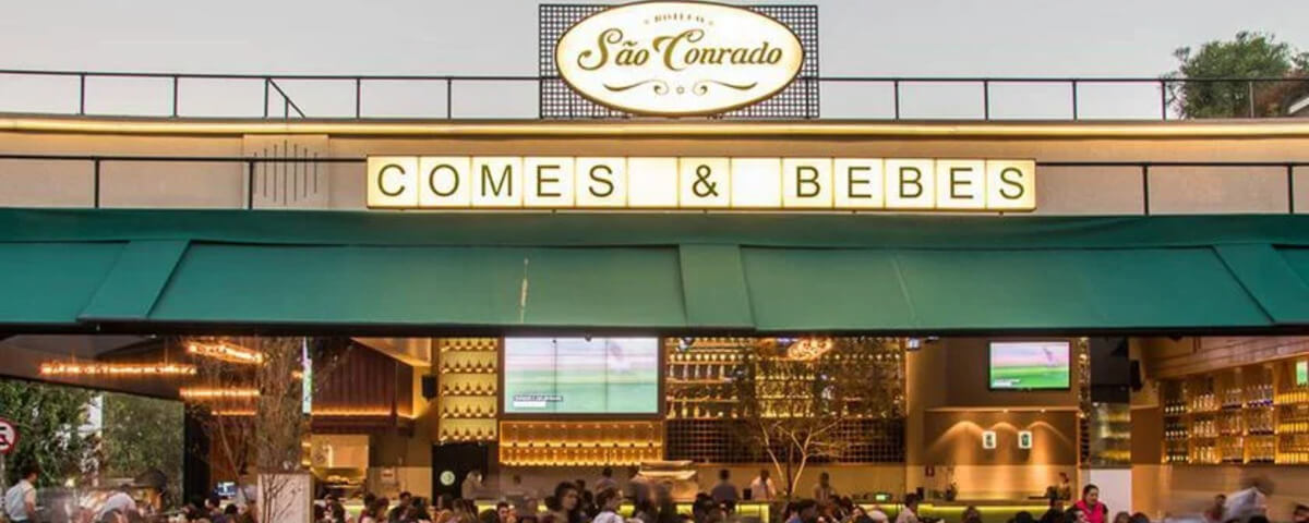 Fachada do Boteco São Conrado, um dos bares para ir em São Paulo, com pessoas sentadas em mesas do lado de fora e televisões passando um jogo de futebol.