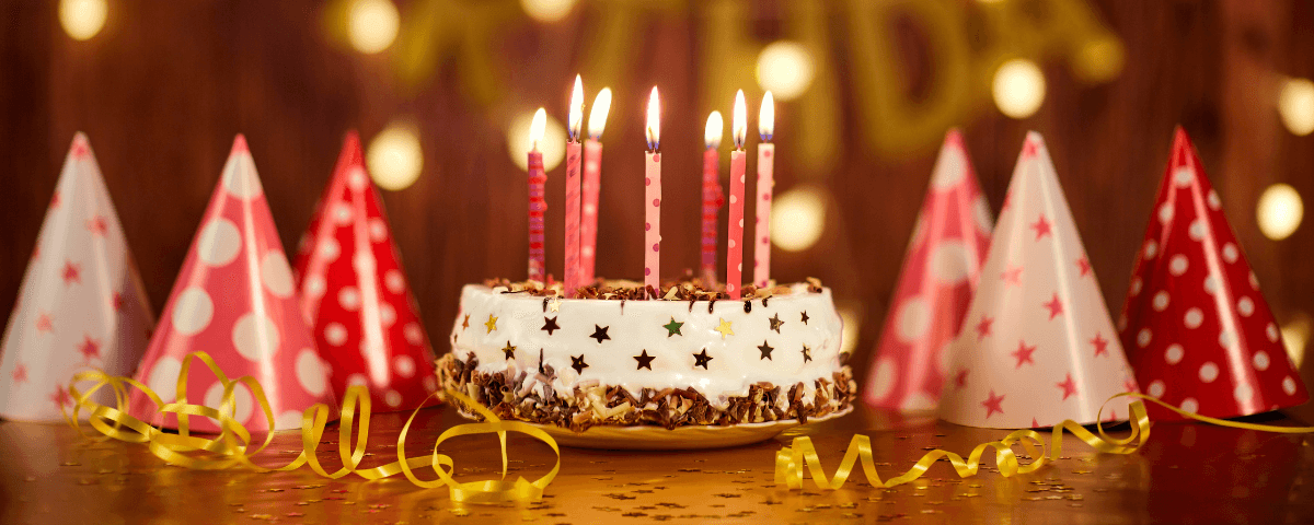 Foto de bolo de aniversário em SP, com velas acesas e chapéus coloridos ao fundo.