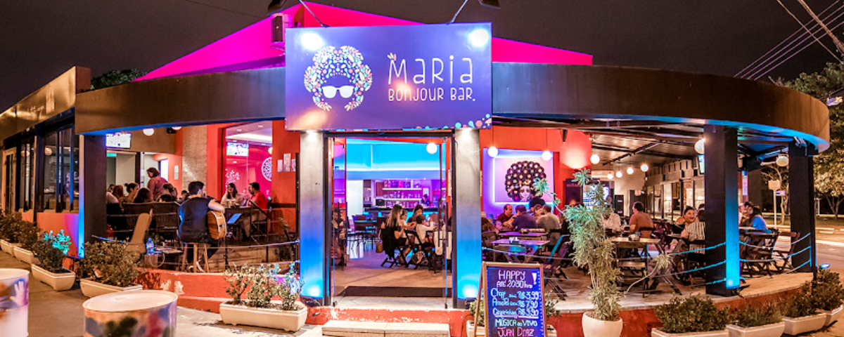 Facahada do Maria Bonjour Bar, com luzes em neon roxa e rosa e com diversas pessoas sentadas, conversando e bebendo. 