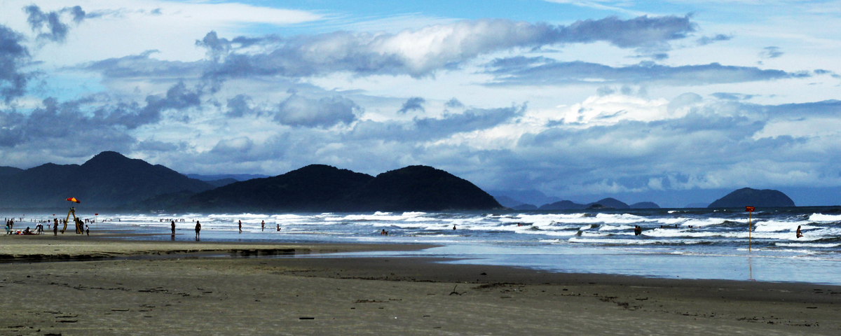 Imagem da praia de Boracéia, com um mar azul que reflete o céu, areia e pessoas caminhando.