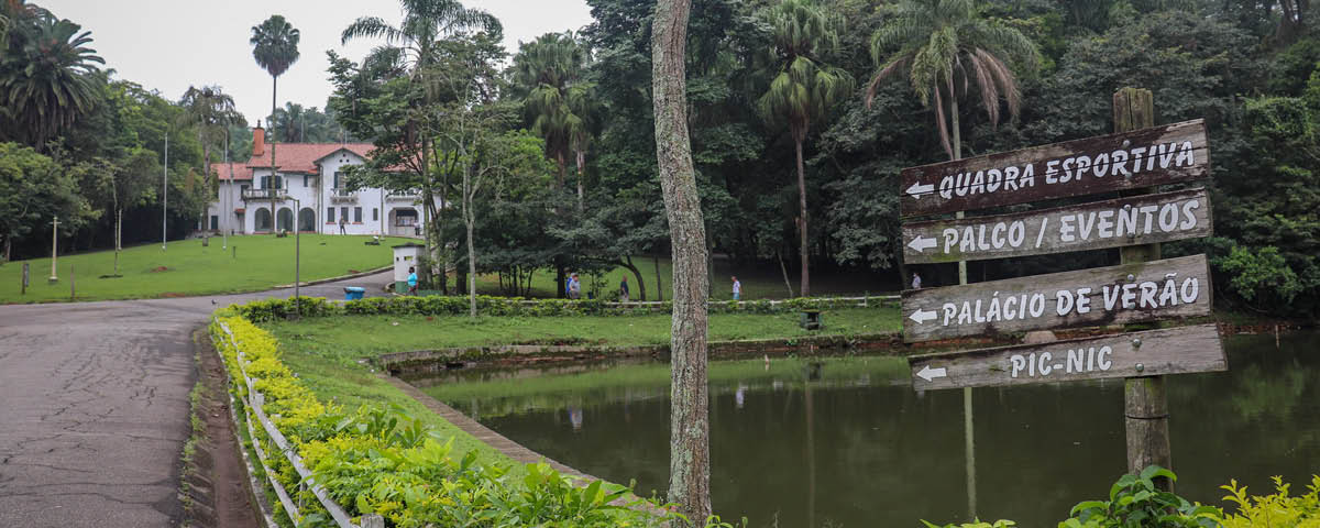Uma das áreas do Horto Florestal, com uma placa indicando direções, árvores, vegetação e uma casa ao fundo.