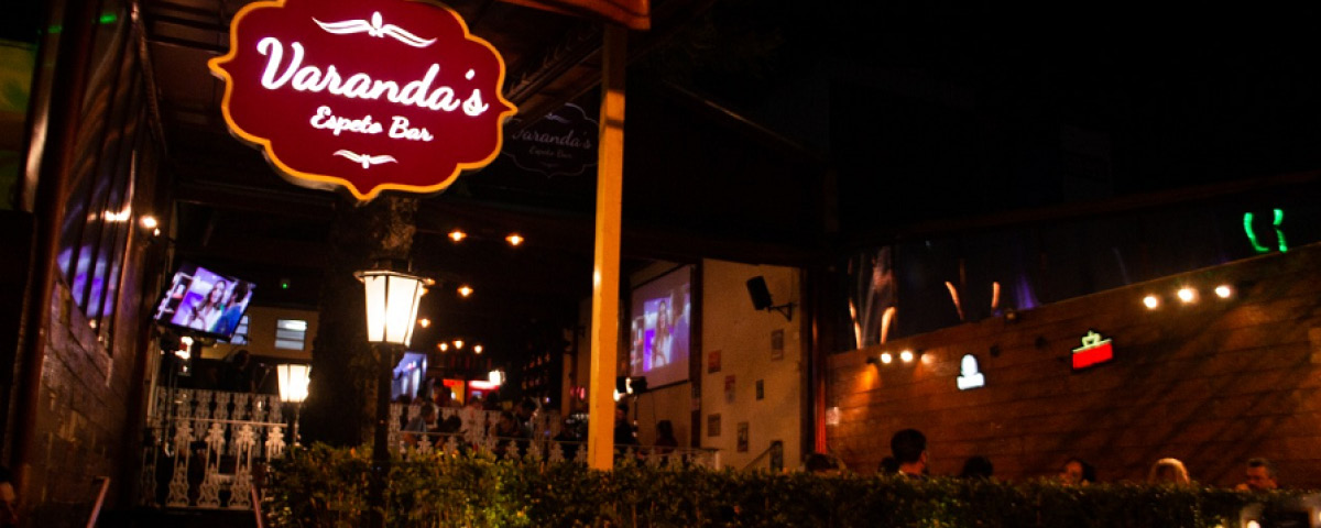 Entrada do Varanda’s Espeto Bar, com uma placa vermelha tendo o seu logotipo e luzes durante a noite.
