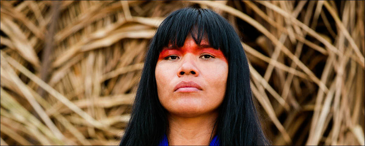 Mulher indígena de cabelo longo escuro com uma feição séria e uma pintura laranja no rosto.