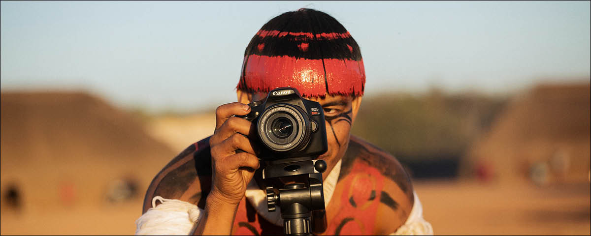 Indígena Xingu tirando foto com uma câmera fotográfica preta.