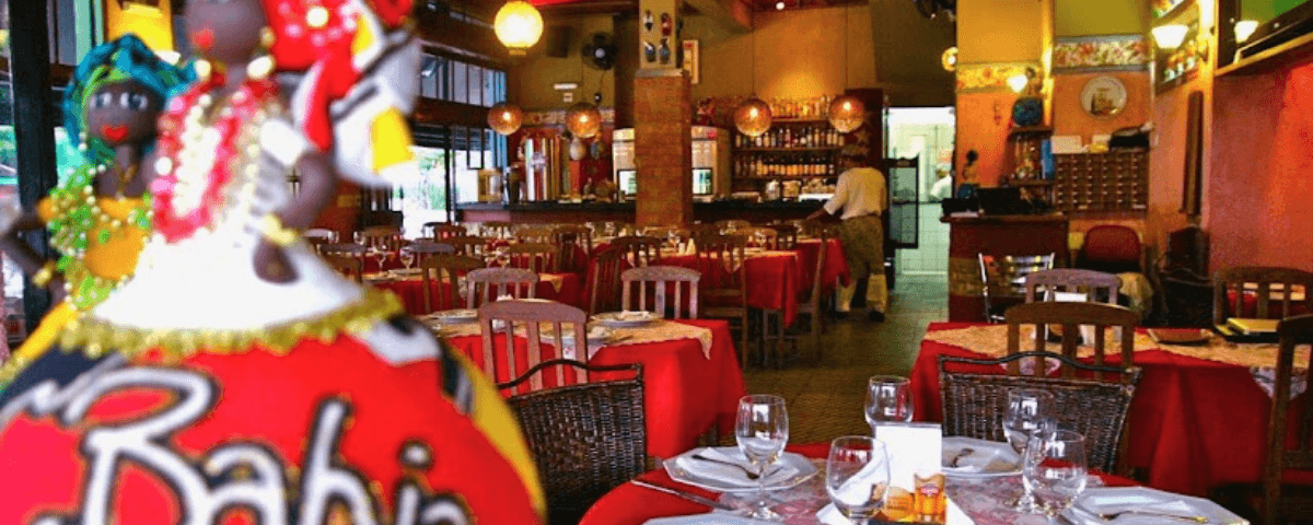 Interior do restaurante Consulado da Bahia, com cadeiras e mesas com toalhas vermelhas, globos de luz e garçom ao fundo da imagem.
