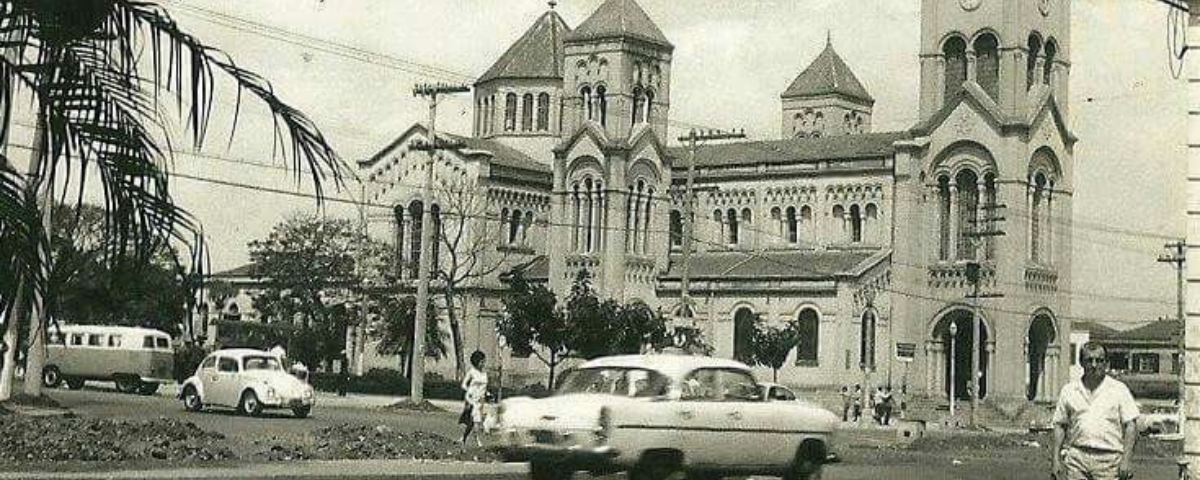 Foto antiga, em preto e branco, do bairro Moema, com sua igreja, pessoas e carros pelas ruas.