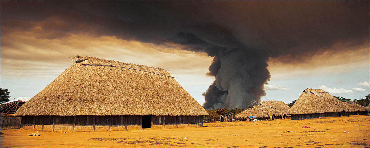 Aldeia indígena, com casinhas de teto de palha e uma fumaça escura ao fundo.