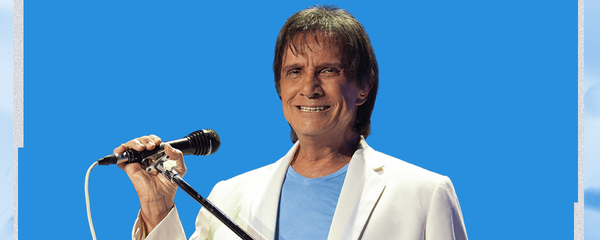 Foto do Roberto Carlos sorrindo para a tela, usando um terno branco, camiseta azul clara num fundo azul. 