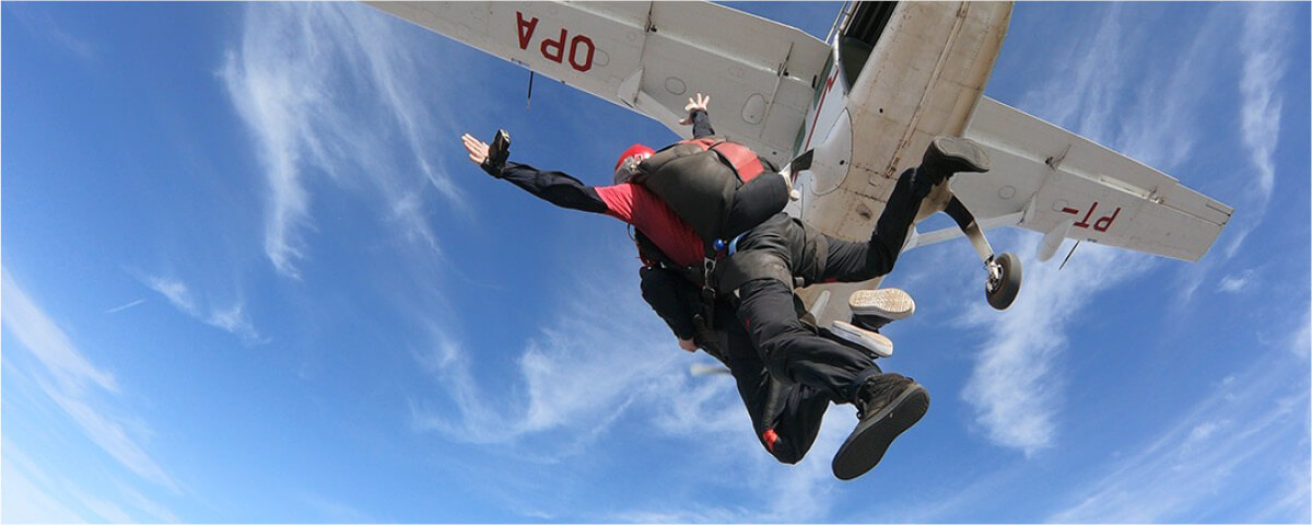 Duas pessoas saltando de paraquedas, com a a visão do céu azul e um avião acima delas.