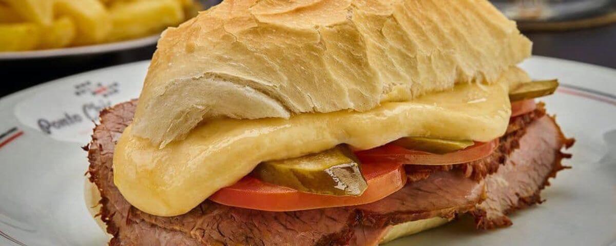 Foto de um dos sanduíches do Ponto Chic com carne, queijo, tomate e picles no pão francês. 