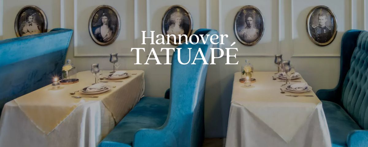 O Hannover Tatuapé tem um banco para casais azuis, quadros na parede e a mesa é composta de uma toalha branca com louças finas em cima. 