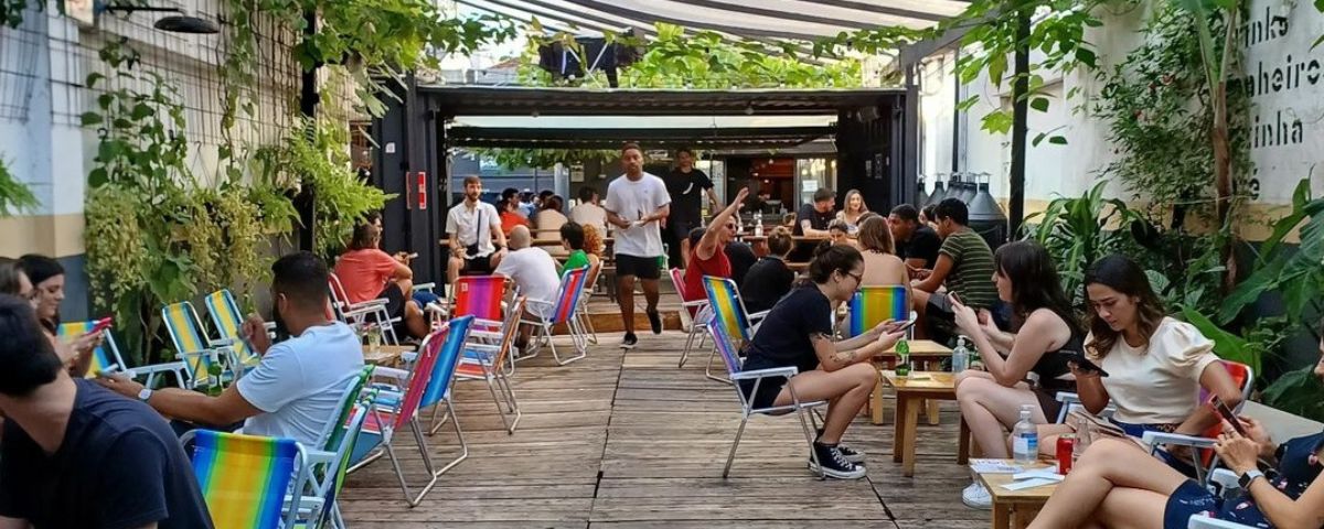 Um dos bares em Pinheiros, o Pitico, tem cadeiras de praia coloridas em ambiente ao livre com pessoas bebendo cervejas e drinks. 