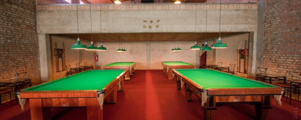 O Atlanta Snooker Bar é um dos maiores bares de sinuca em SP. Quatro mesas de sinuca com luminárias em cima estão em uma sala que tem parede de tijolos. 
