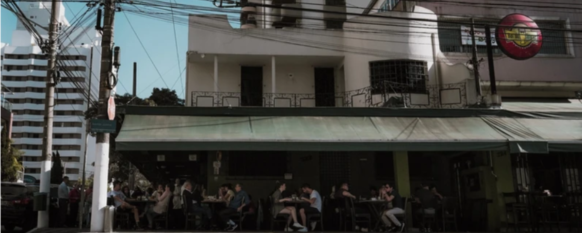 Uma das opções para almoço em São Paulo é o Bar do Juarez, que tem um toldo verde em sua varanda que fica em uma esquina. O bar tem várias pessoas sentadas nas mesas. 