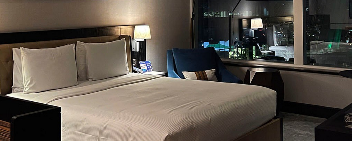 O quarto do Hotel Hilton Morumbi tem uma cama de casal com roupa de cama branca, mesa de cabeceira com abajour e uma poltrona azul do lado direito. 