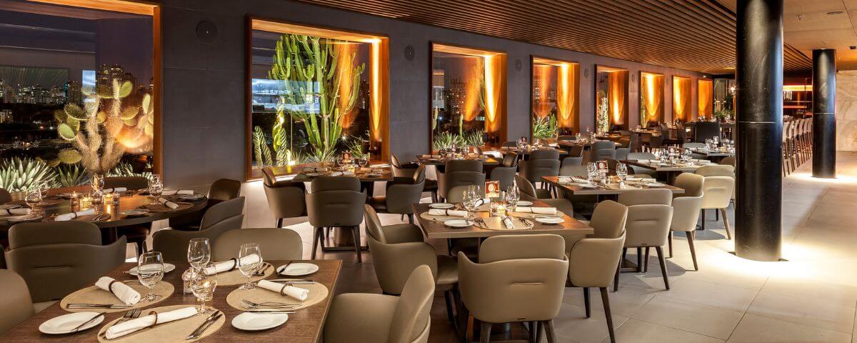 Uma incrível opção para o Dia dos Namorados em SP é o restaurante Skye. O salão é elegante, com cadeiras beges e mesas de madeiras. Ao longo do salão tem duas pilastras pretas e janelas grandes. 