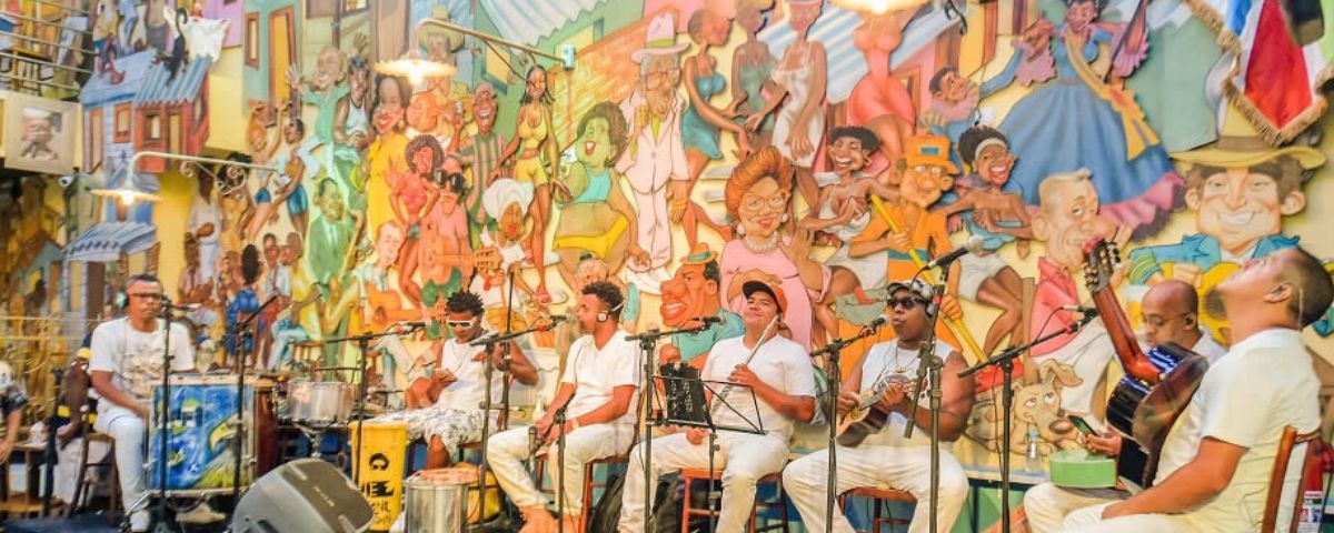 Banda toca samba em São Paulo. Ao fundo um mural com diversos desenhos de figuras conhecidas do samba. 