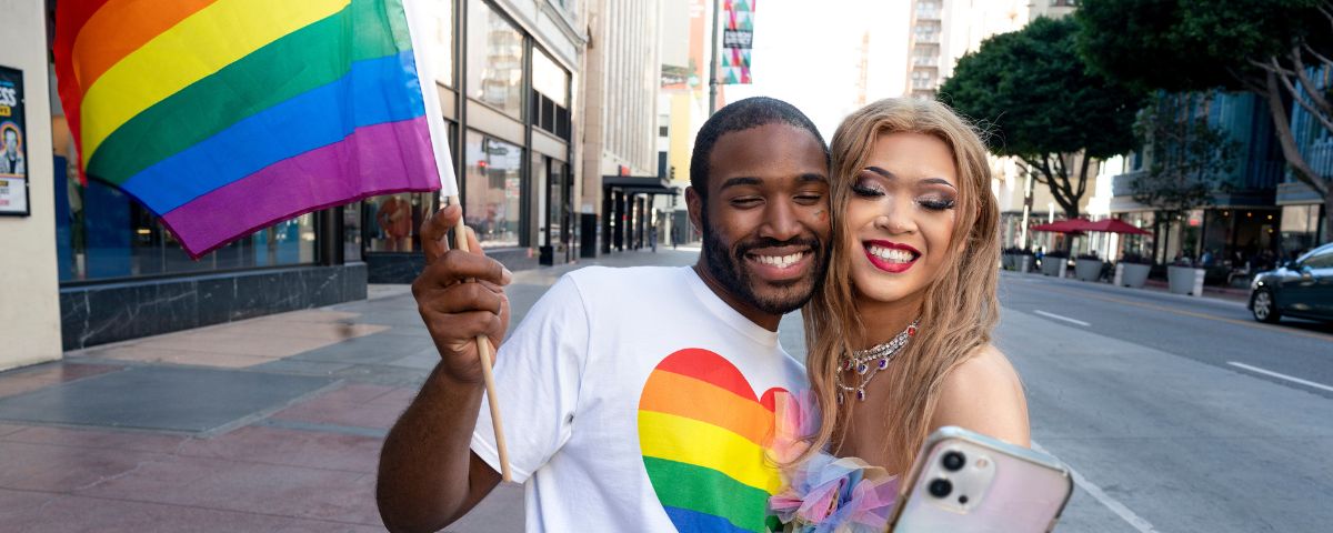 Na Parada do Orgulho LGBT+ de São Paulo algumas figuras importantes do movimento contra o preconceito serão homenageadas. Dois amigos tiram selfie, um deles segura uma bandeira colorida e usa uma blusa com um coração no meio, com as cores da bandeira. 