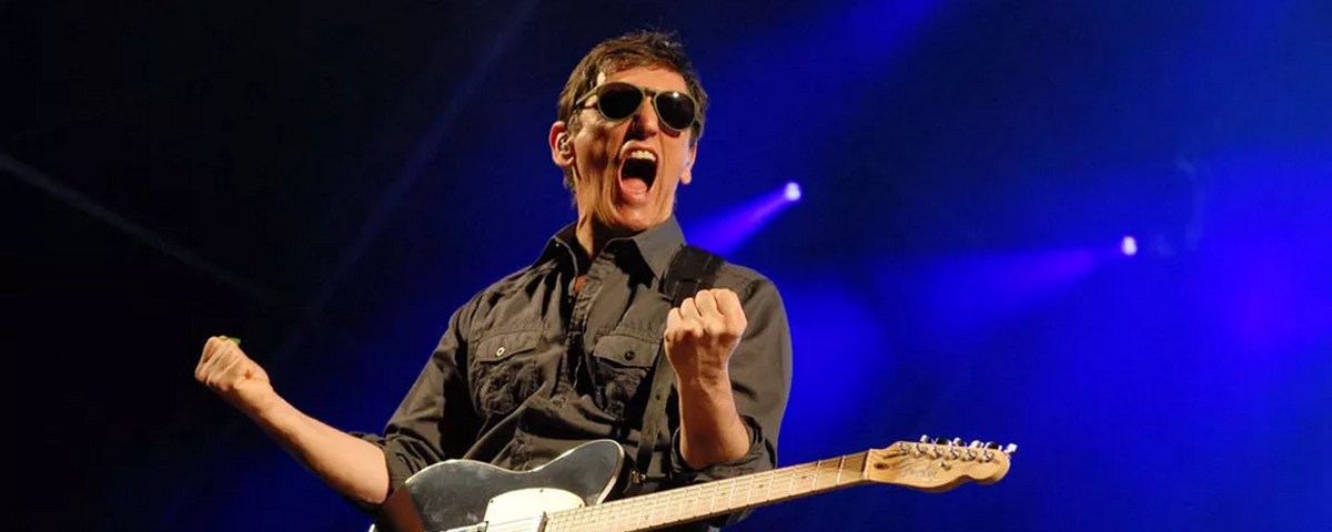O cantor Alexandre Durão vibra no palco usando óculos escuros e com uma guitarra pendurada no pescoço. O artista se apresenta no Centro Histórico de São Paulo para celebrar o Dia Mundial do Rock. 