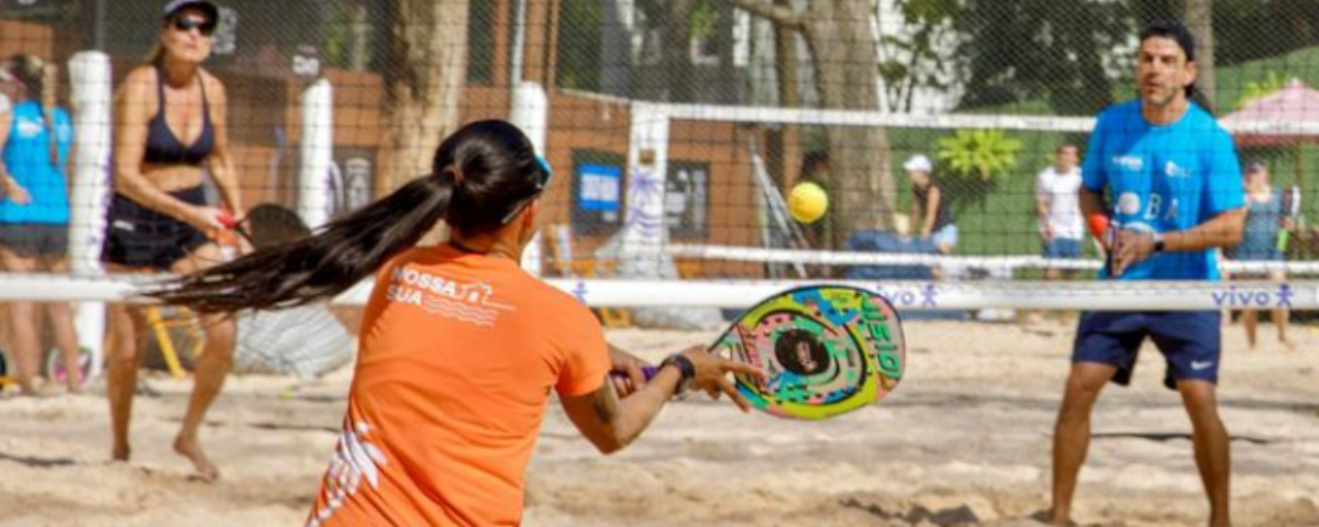 Duplas se enfrentam na quadra de areia, com uma rede no centro. Mulher de blusa laranja segura a raquete enquanto vai com ela em direção a bola. 