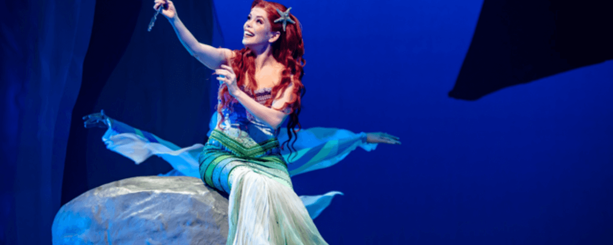 Atriz representando a Ariel do conto "A Pequena Sereia" está sentada em uma pedra segurando um pequeno espelho, por onde ela olha o seus cabelos vermelhos enquanto sorri. 